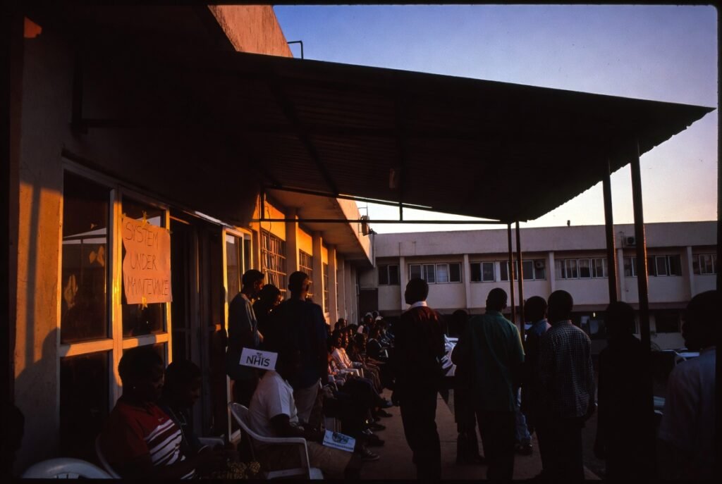 Patients queue outside Kumasi government hospital at dawn, NHIS card visible, system under maintenance sign on door